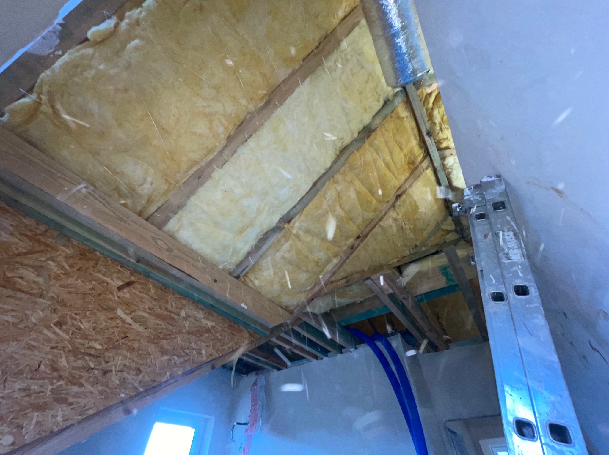 Poddasze w trakcie ocieplania wełną mineralną, widoczne krokwie, płyta OSB i rury instalacyjne, drabina oparta o ścianę.