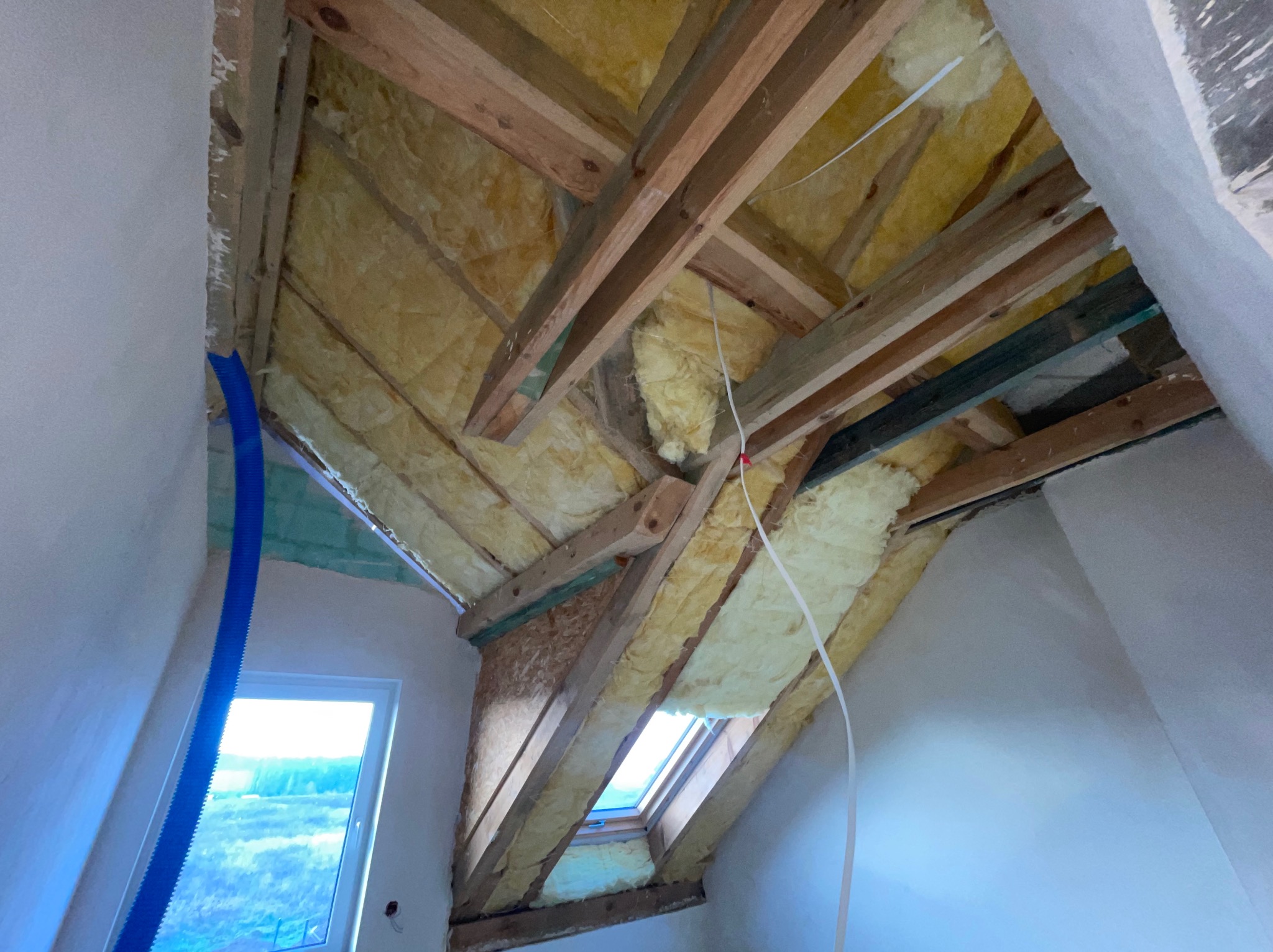 Poddasze w trakcie ocieplania wełną mineralną, widoczne drewniane krokwie, okno dachowe i fragment nieotynkowanej ściany z niebieskim przewodem wentylacyjnym.