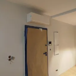 Biała klimatyzacja na ścianie nad zabezpieczonymi drzwiami w trakcie remontu. Widoczne kable elektryczne i skrzynka bezpieczników.