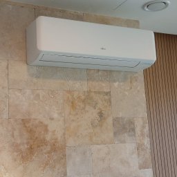 Cool Fox Sp. z o.o. - Biała klimatyzacja Fujitsu na ścianie z trawertynu, obok pionowe panele drewniane. Widoczny fragment skrzynki elektrycznej po lewej stronie.