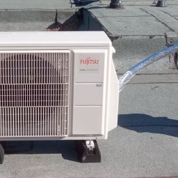 Cool Fox Sp. z o.o. - Biała jednostka zewnętrzna klimatyzacji Fujitsu na dachu z szarą papą. Widoczny srebrny przewód odprowadzający powietrze. Montaż na czarnych podkładkach antywibracyjnych.
