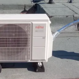 Biała jednostka zewnętrzna klimatyzacji Fujitsu na dachu z szarą papą. Widoczny srebrny przewód odprowadzający powietrze. Montaż na czarnych podkładkach antywibracyjnych.