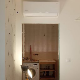 Cool Fox Sp. z o.o. - Biały klimatyzator na ścianie z wzorzystą tapetą, widok na łazienkę w trakcie remontu z pralką i lampą budowlaną.