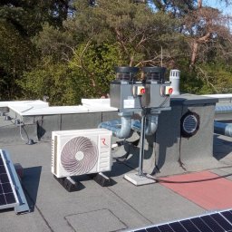 Cool Fox Sp. z o.o. - Instalacja klimatyzacji Rotenso na dachu budynku z panelami słonecznymi, widoczne przewody wentylacyjne i komin. Tło stanowią drzewa i błękitne niebo.
