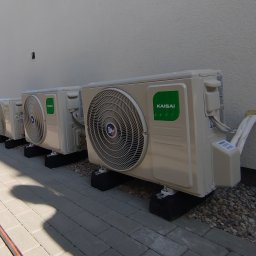 Cool Fox Sp. z o.o. - Trzy klimatyzatory Kaisai zamontowane na zewnątrz budynku, widoczne przewody i instalacja. Biała ściana i szara kostka brukowa. Częściowo widoczny samochód dostawczy.
