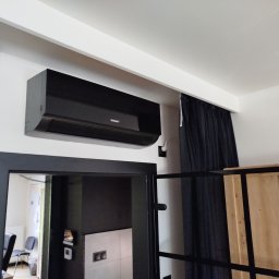 Cool Fox Sp. z o.o. - Czarny klimatyzator ścienny marki Kaisai zamontowany w pomieszczeniu z czarną stolarką drzwiową i zasłonami. Widoczny fragment wnętrza biura.