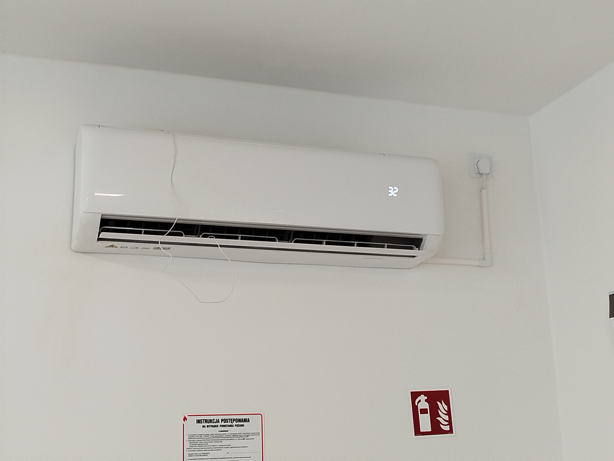 Biały klimatyzator AUX z wyświetlaczem 32°C, zamontowany na ścianie w Sianowskiej Hucie. Widoczna instrukcja ppoż. i instalacja elektryczna.