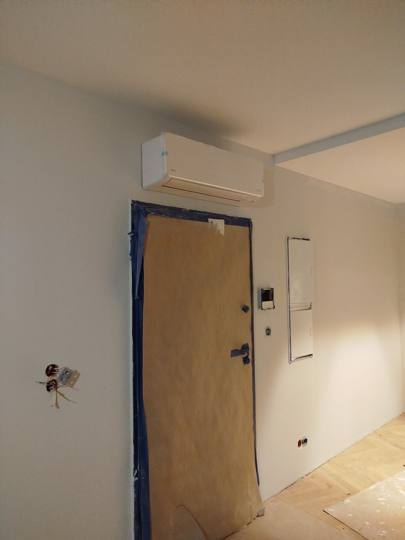 Biała klimatyzacja na ścianie nad zabezpieczonymi drzwiami w trakcie remontu. Widoczne kable elektryczne i skrzynka bezpieczników.