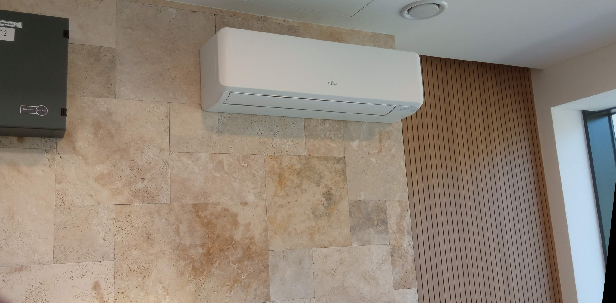 Biała klimatyzacja Fujitsu na ścianie z trawertynu, obok pionowe panele drewniane. Widoczny fragment skrzynki elektrycznej po lewej stronie.