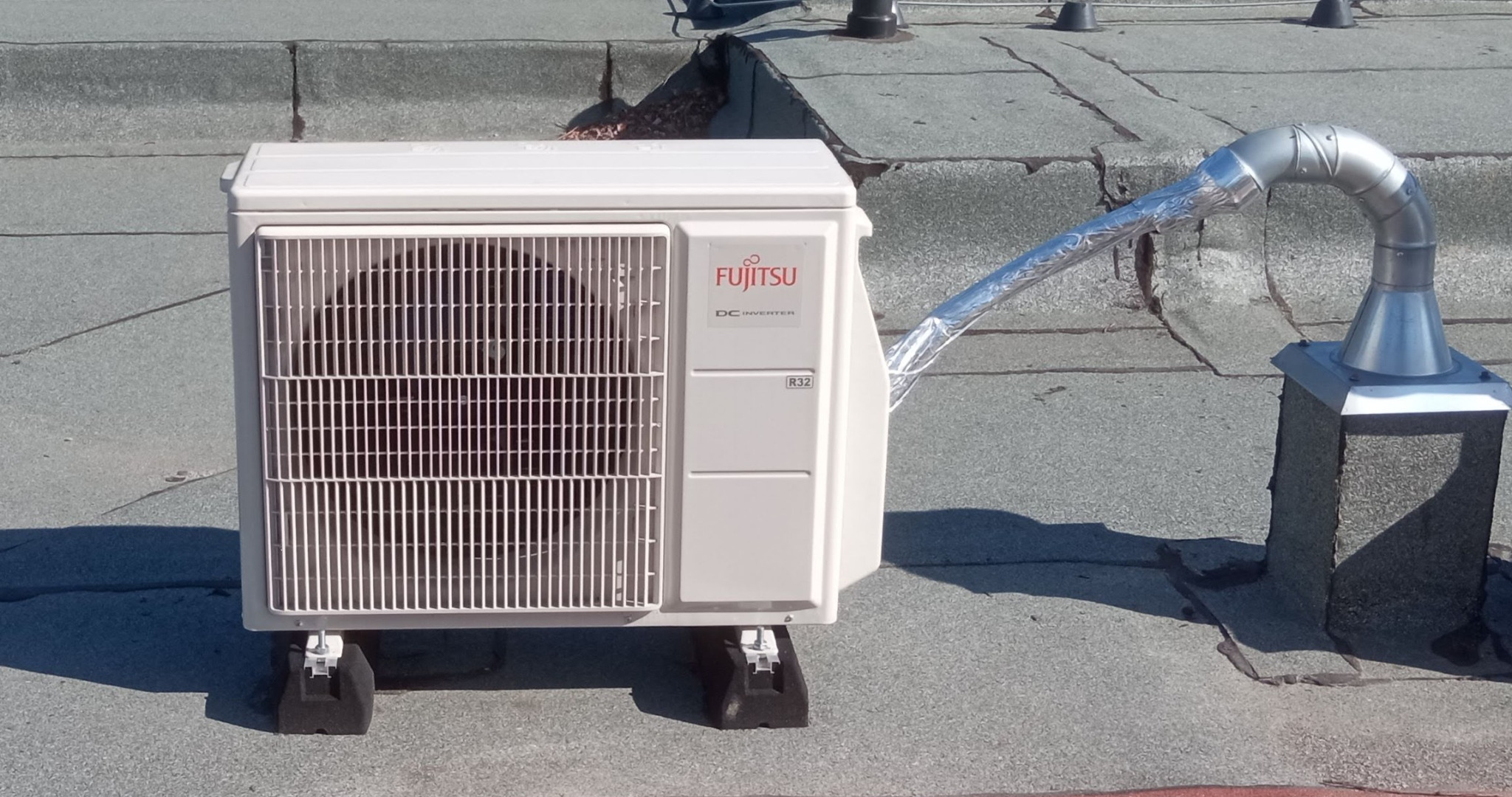 Biała jednostka zewnętrzna klimatyzacji Fujitsu na dachu z szarą papą. Widoczny srebrny przewód odprowadzający powietrze. Montaż na czarnych podkładkach antywibracyjnych.