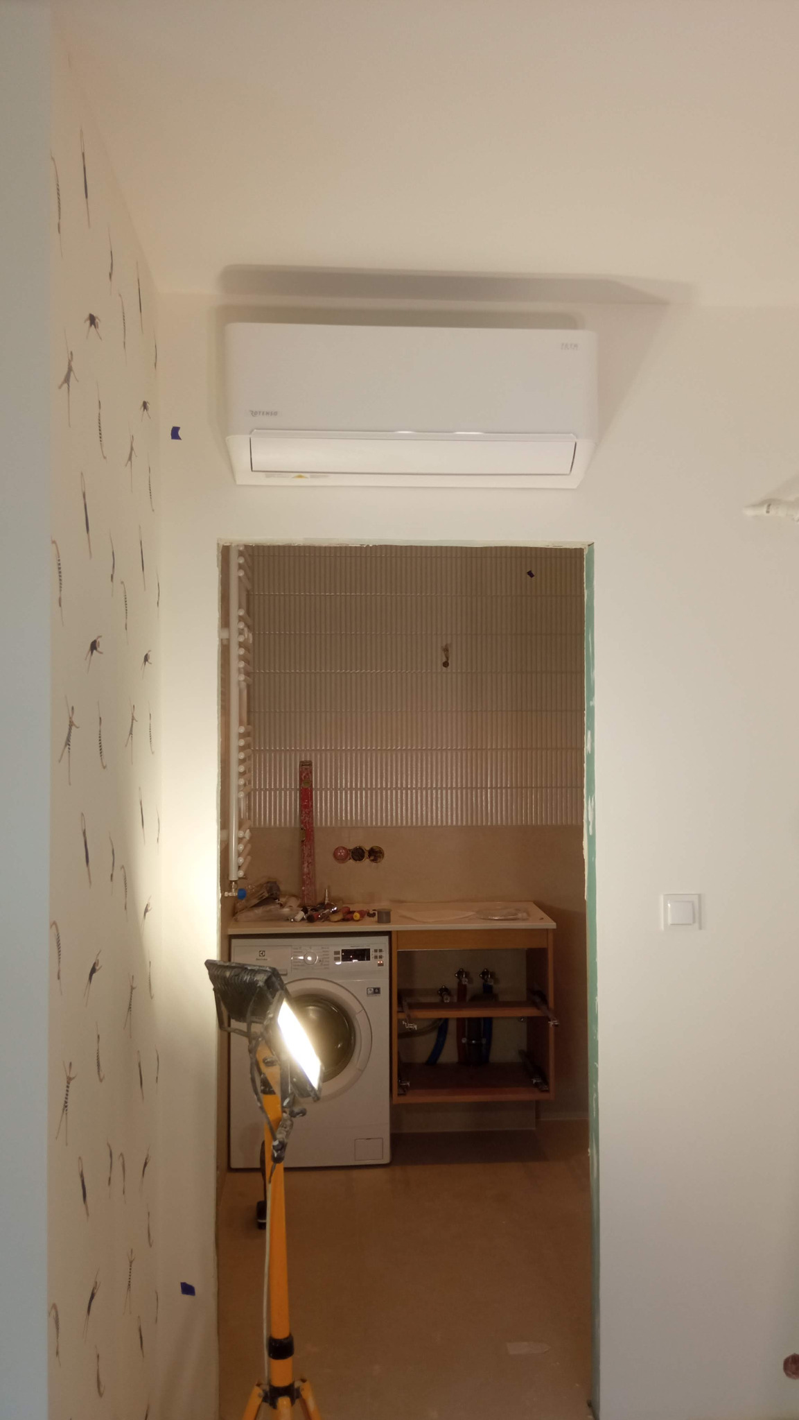 Biały klimatyzator na ścianie z wzorzystą tapetą, widok na łazienkę w trakcie remontu z pralką i lampą budowlaną.