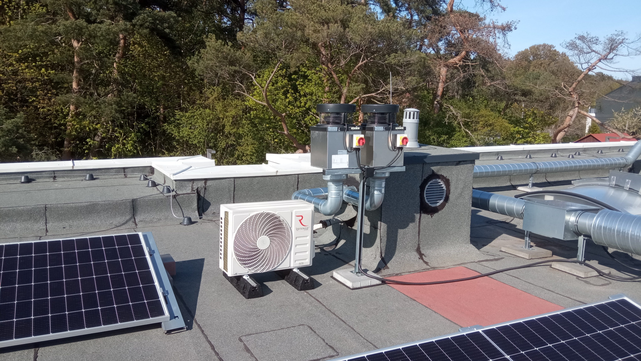 Instalacja klimatyzacji Rotenso na dachu budynku z panelami słonecznymi, widoczne przewody wentylacyjne i komin. Tło stanowią drzewa i błękitne niebo.