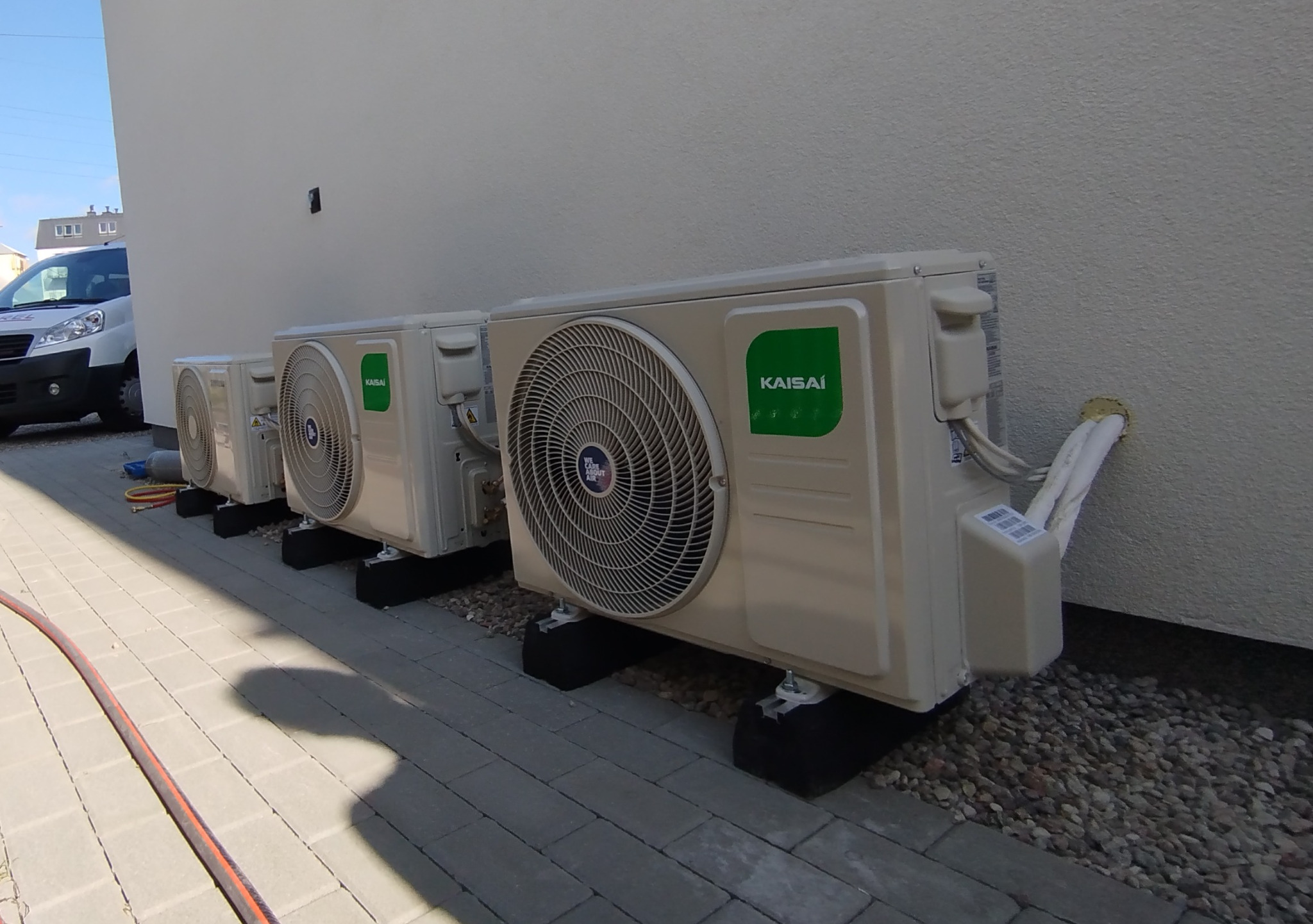 Trzy klimatyzatory Kaisai zamontowane na zewnątrz budynku, widoczne przewody i instalacja. Biała ściana i szara kostka brukowa. Częściowo widoczny samochód dostawczy.