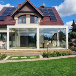Prim - Szklana zabudowa tarasu przylegająca do domu z panelami słonecznymi na dachu, widok z trawnika.