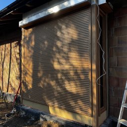 Prim - Zewnętrzna roleta okienna w drewnianej ramie, zamontowana w budynku z cegły. Widoczne przewody elektryczne i drabina, sugerujące trwający montaż.