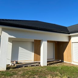 Prim - Biały dom z roletami zewnętrznymi w kolorze białym i drewnianą elewacją. Widoczne puszki z farbą na zewnątrz. Błękitne niebo w tle, trawa. Ujęcie z dołu.