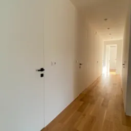 Nowoczesny korytarz z minimalistycznymi, białymi drzwiami wewnętrznymi z czarnymi klamkami i drewnianą podłogą. Widok w głąb, wpadające światło dzienne.