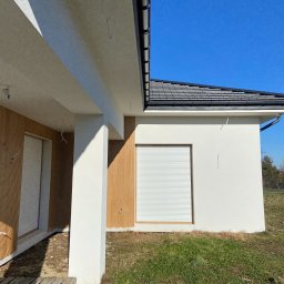 Prim - Nowoczesny dom z drewnianą elewacją i roletami zewnętrznymi w kolorze białym. Widok na fragment budynku z ogrodem w słoneczny dzień. Elementy wykończeniowe w kolorze brązowym.