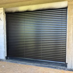 Prim - Zamknięte, poziome żaluzje w kolorze grafitowym, zamontowane w surowym otworze okiennym w trakcie budowy. Widoczne bloczki i surowy strop.