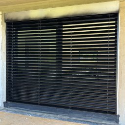 Prim - Zamknięte, czarne żaluzje fasadowe w surowym otoczeniu budowlanym. Widoczne niedoskonałości tynku i fragment podłogi z płyty OSB. Minimalistyczny design.