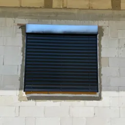 Czarne żaluzje fasadowe zamontowane w oknie budynku w trakcie budowy, widoczne białe bloczki i drewniane elementy konstrukcji dachu.