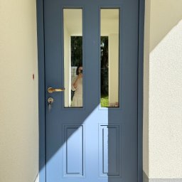 Prim - Błękitne drzwi zewnętrzne z dwoma przeszkleniami, widoczne klucze w zamku, oświetlone promieniami słońca, z odbiciem osoby robiącej zdjęcie.