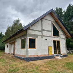 Prim - Surowy dom z pustaków z zamontowanymi oknami o drewnianych ramach i ciemnym dachem, na tle drzew i pochmurnego nieba. Widoczna żółta tablica informacyjna.