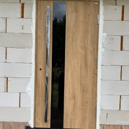 Prim - Zamontowane drzwi zewnętrzne z drewnopodobnym wykończeniem i pionowym, czarnym przeszkleniem, osadzone w murze z betonu komórkowego. Częściowo zabezpieczona folią ochronną.