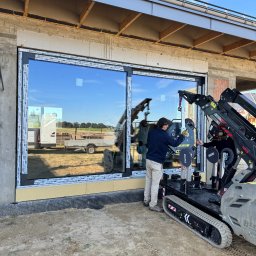 Prim - Montaż okna PCV przez dwóch pracowników z użyciem specjalistycznego podnośnika gąsienicowego. Widoczne okno z ramą Okoplast, budynek w trakcie budowy.