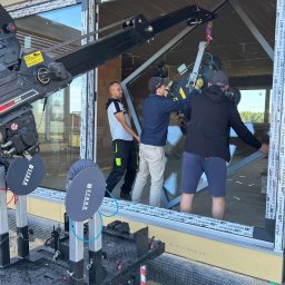 Prim - Montaż dużego okna z użyciem dźwigu BEFARD. Trzech monterów w trakcie instalacji okna w surowym budynku. Widoczne detale konstrukcyjne i narzędzia.