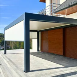 Nowoczesna, grafitowa pergola z roletą zewnętrzną i drewnianą ścianą w tle, na tle błękitnego nieba. Widoczna kostka brukowa i fragment budynku.
