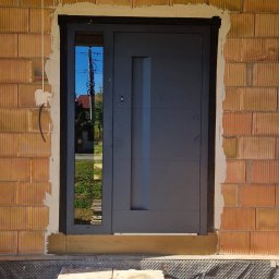 Prim - Montaż antracytowych drzwi zewnętrznych z szybą w Dębicy. Widoczny surowy mur z cegły i tymczasowe schody z płyt betonowych. Nowoczesny design.