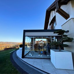 Prim - Nowoczesna, przeszklona zabudowa balkonu z czarną ramą, widok z zewnątrz na tle jesiennego krajobrazu. Minimalistyczny design, drzewko bonsai w donicy.