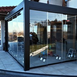 Prim - Szklana zabudowa tarasu z czarną ramą w trakcie montażu. Widoczne narzędzia i drabina. W tle dom z czerwonym dachem i basen.