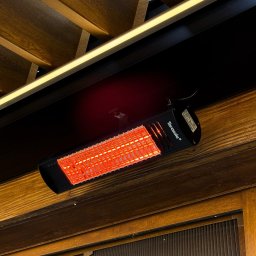 Prim - Czarny promiennik ciepła Tarasola zamontowany pod drewnianym sufitem z oświetleniem LED, widoczny fragment drewnianej ściany z żaluzjami.