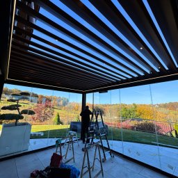 Prim - Nowoczesna zabudowa balkonu z przesuwnymi szklanymi panelami i drewnianą pergolą z oświetleniem LED. Widok na jesienny ogród. Prace montażowe w toku.