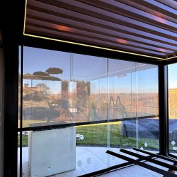 Prim - Nowoczesna zabudowa tarasu z przesuwnymi panelami szklanymi i roletami screen, widok na ogród. Oświetlenie LED podkreśla drewnianą konstrukcję pergoli.