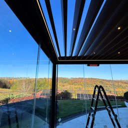 Prim - Nowoczesna zabudowa tarasu z czarną pergolą lamelową i szklanymi ścianami, widok na jesienny krajobraz. Drabina oparta o ścianę.