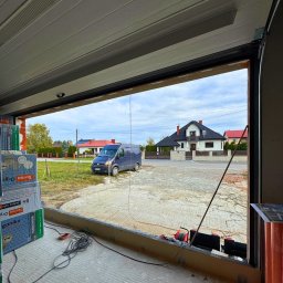 Prim - Montaż bramy garażowej segmentowej: widok z wnętrza garażu na podjazd i dom. Widoczne narzędzia i materiały budowlane. Brama w trakcie instalacji.