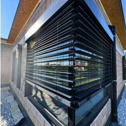 Prim - Nowoczesne, czarne żaluzje fasadowe zamontowane na oknie budynku w trakcie budowy, widoczne cegły i elementy konstrukcyjne. Częściowo opuszczone lamele.