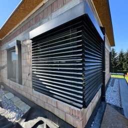 Prim - Zamontowane czarne żaluzje fasadowe na oknie budynku w trakcie budowy, widoczne białe kamienie i folia budowlana. Ujęcie z dołu, błękitne niebo.