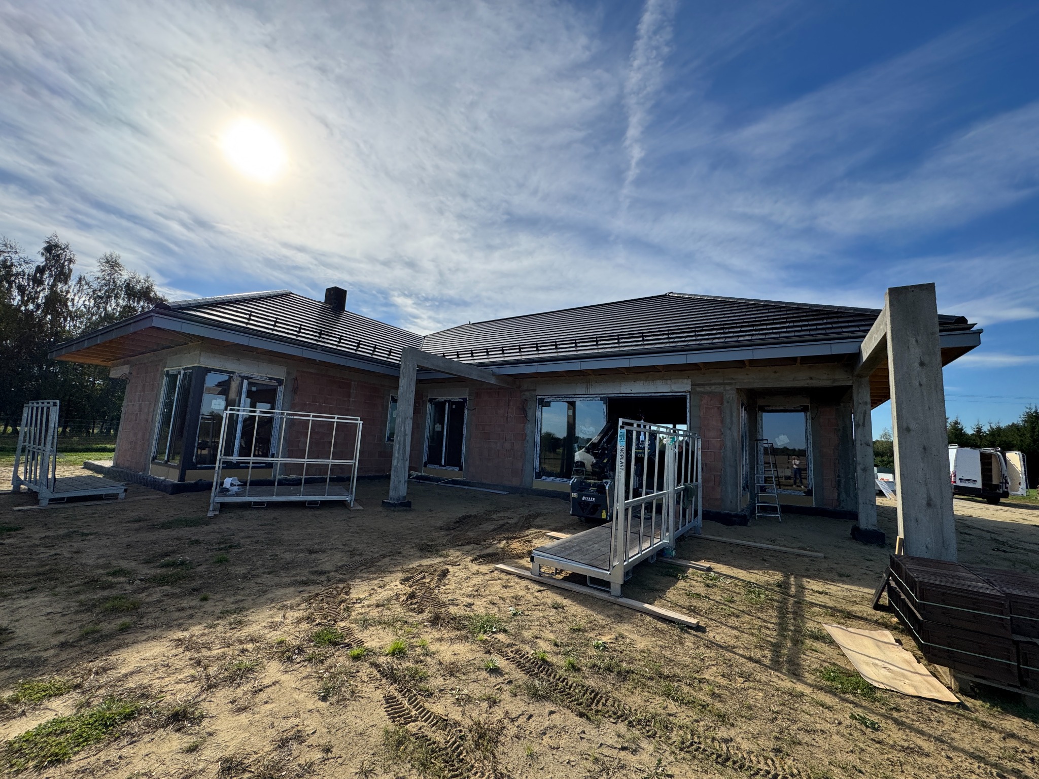 Dom w trakcie budowy z widocznymi oknami i elementami konstrukcyjnymi. Na pierwszym planie podesty robocze i materiały budowlane. Słoneczny dzień, błękitne niebo.