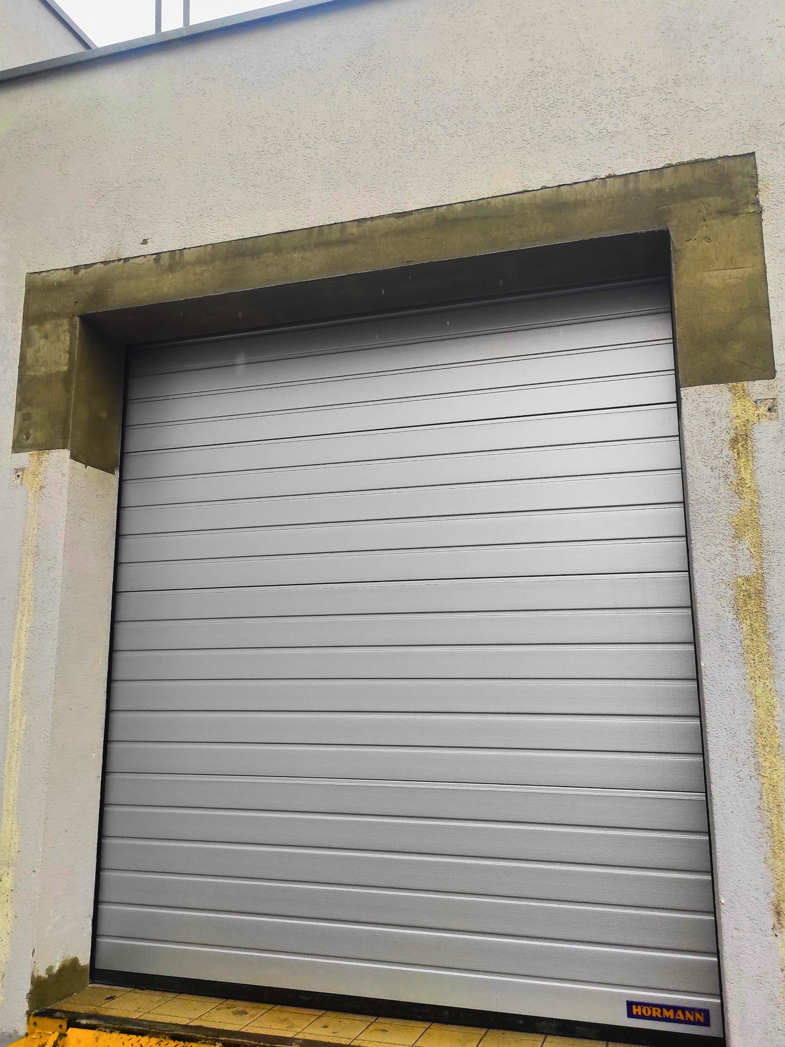Zamknięta, szara brama garażowa segmentowa marki Hörmann, widoczna z bliska, z żółtym najazdem i betonową obudową. Na dole widoczne logo producenta.