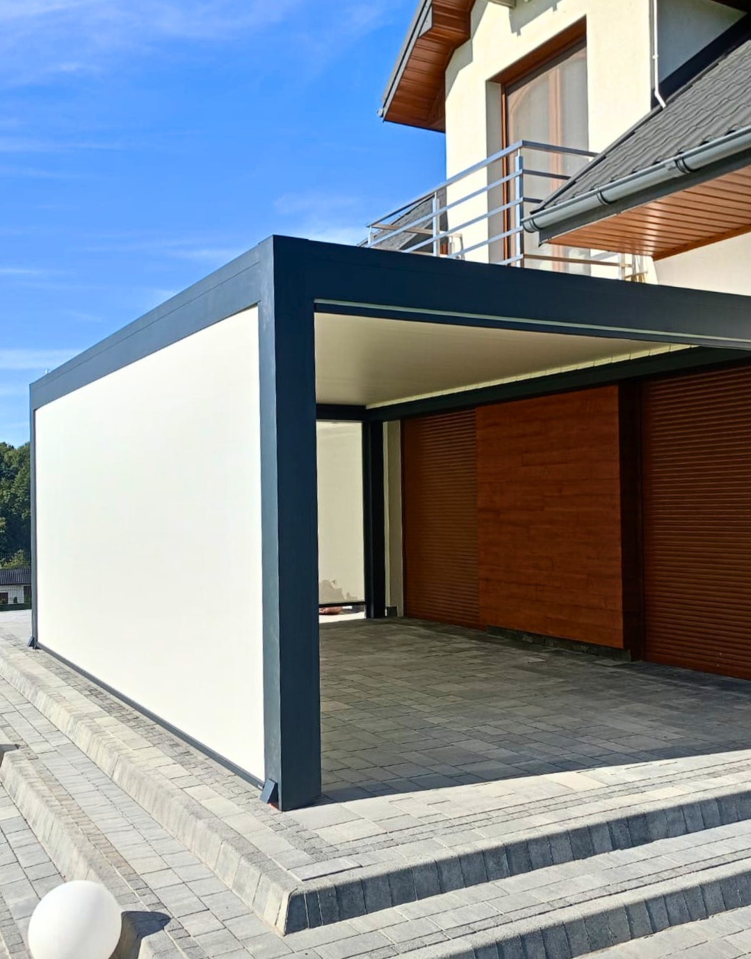 Nowoczesna pergola z przesuwnymi panelami drewnianymi i białą ścianą boczną, stanowiąca zadaszenie tarasu przy domu, widok z dołu na tle błękitnego nieba.