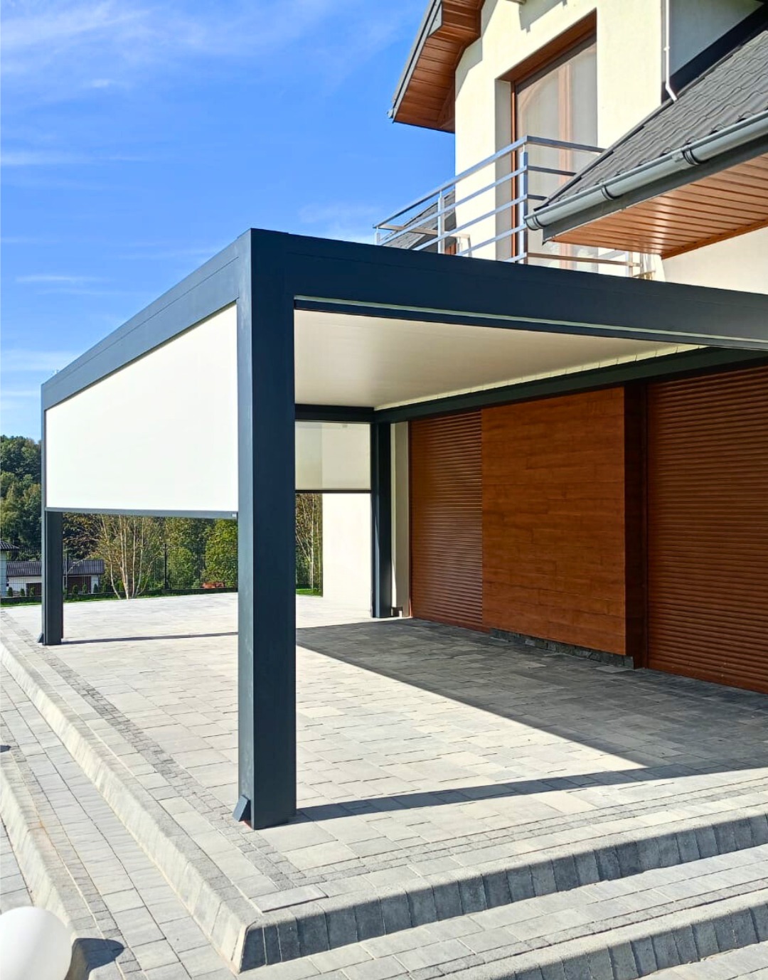 Nowoczesna, grafitowa pergola z roletą zewnętrzną i drewnianą ścianą w tle, na tle błękitnego nieba. Widoczna kostka brukowa i fragment budynku.