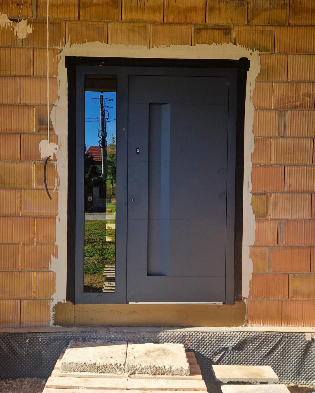 Montaż antracytowych drzwi zewnętrznych z szybą w Dębicy. Widoczny surowy mur z cegły i tymczasowe schody z płyt betonowych. Nowoczesny design.