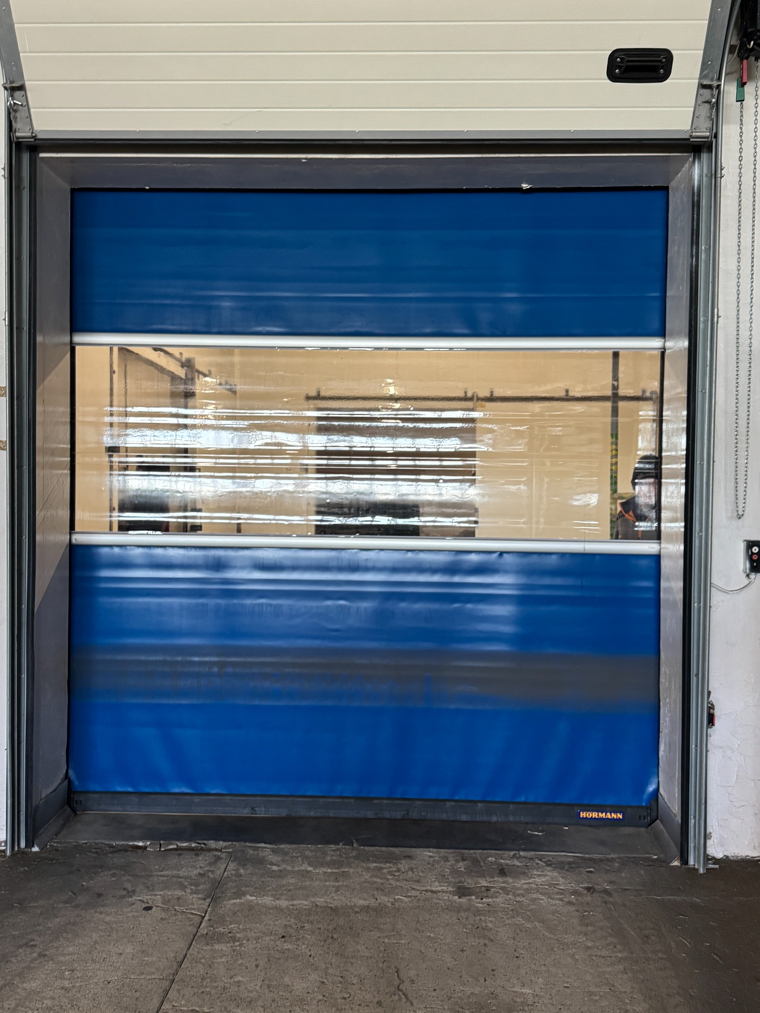 Niebieska, segmentowa brama garażowa z oknem na środku, widoczna od wewnątrz. Brama częściowo otwarta, z widocznym logo Hormann na dole.