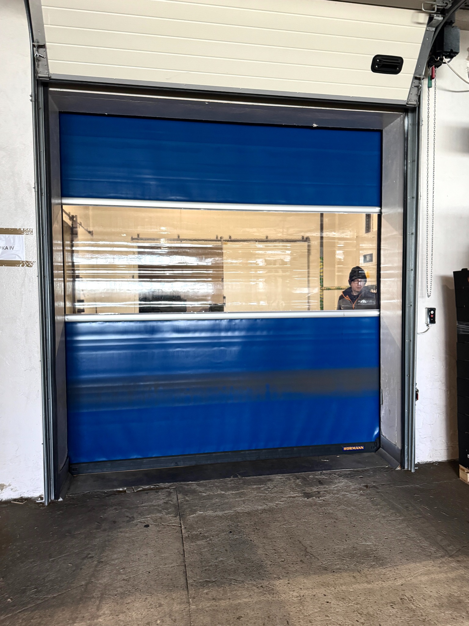 Niebieska przemysłowa brama segmentowa z oknem, przez które widać sylwetkę osoby; brama marki Hormann, zamontowana w hali.