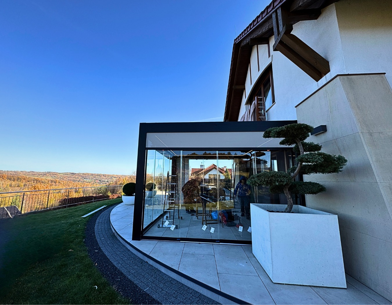 Nowoczesna, przeszklona zabudowa balkonu z czarną ramą, widok z zewnątrz na tle jesiennego krajobrazu. Minimalistyczny design, drzewko bonsai w donicy.