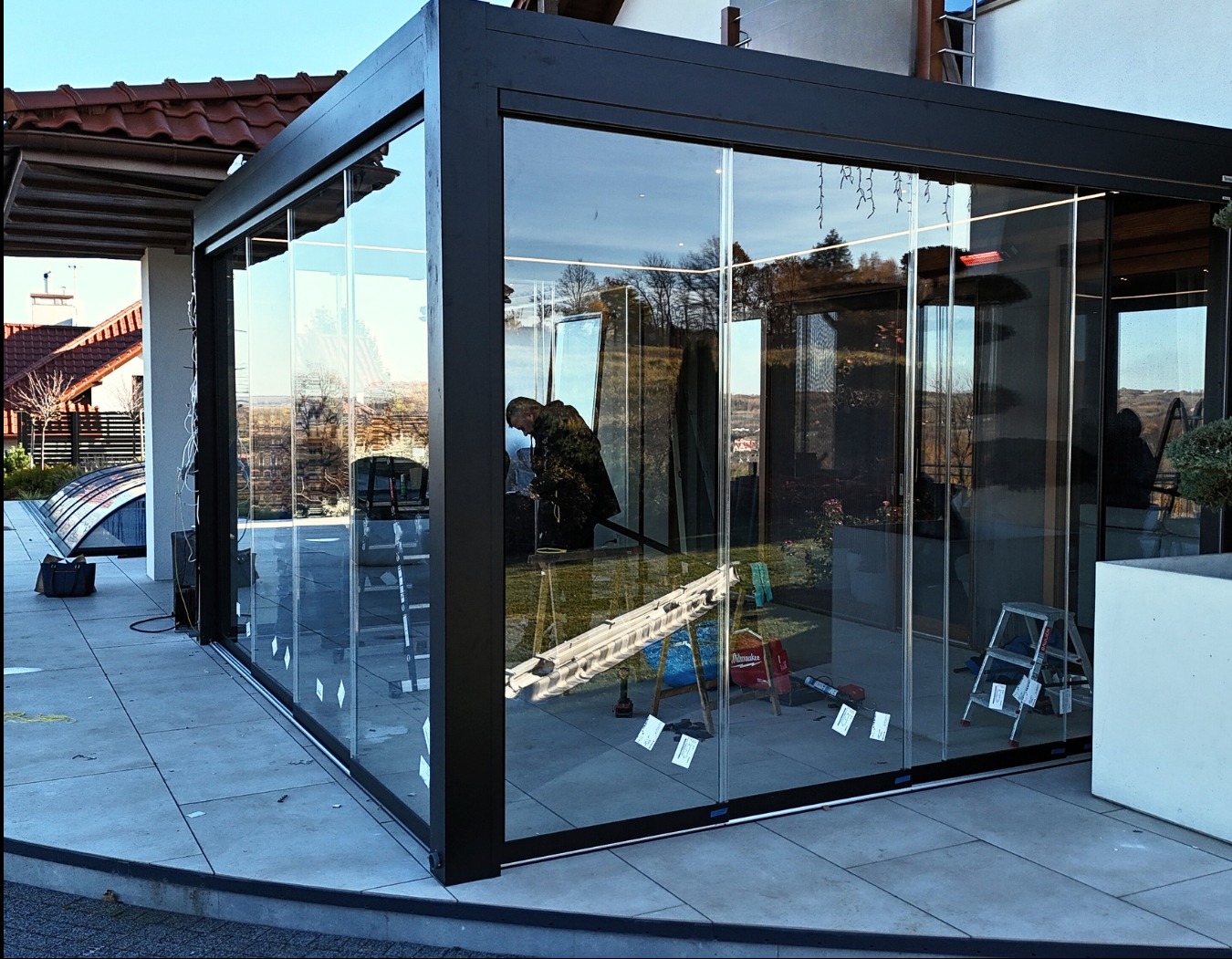 Szklana zabudowa tarasu z czarną ramą w trakcie montażu. Widoczne narzędzia i drabina. W tle dom z czerwonym dachem i basen.